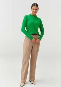 Groene lange mouwen top met een hoge halslijn, gecombineerd met beige high-waisted broek. Schoenen hebben een puntige neus en zwarte hakaccenten.