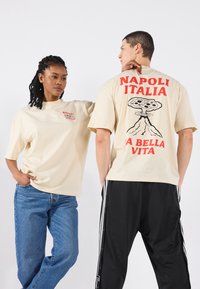 Beige bomulls t-skjorter med røde grafikker: "NAPOLI ITALIA" og et pizzasymbol, båret av to modeller med forskjellige denimstiler.