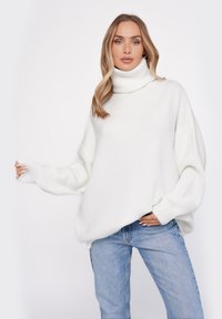 Biały, oversize'owy sweter z golfem o prążkowanej fakturze, z obniżonymi ramionami i szerokimi rękawami, w połączeniu z jasnoniebieskimi dżinsami.