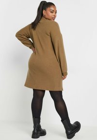 Braunes, geripptes Strickkleid mit langen Ärmeln, bis zur Mitte des Oberschenkels fallend, kombiniert mit schwarzen Knöchelstiefeln und transparenten schwarzen Strumpfhosen.