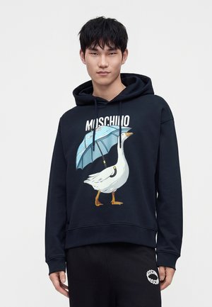 Noor mees musta kapuutsiga pusa seljas, valge hane hoides sinist vihmavarju ning Moschino brändinimi graafika kohal.
