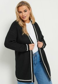 Svart stickad kofta med vit kant, öppen front, långa ärmar, texturerad tyg; bärs över en vit skjorta och blå jeans.