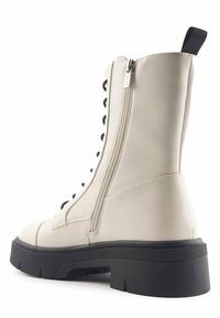 Botín de tobillo de imitación de cuero blanco con cordones negros, cremallera trasera y suela de goma texturizada negra. Presenta costuras decorativas en la parte superior.
