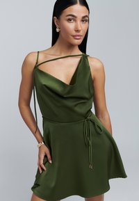 Groene satijnen jurk met een gedrapeerde halslijn, dunne bandjes en een strik rond de taille. Heeft een uitlopende rok en een subtiele glans in de stof.