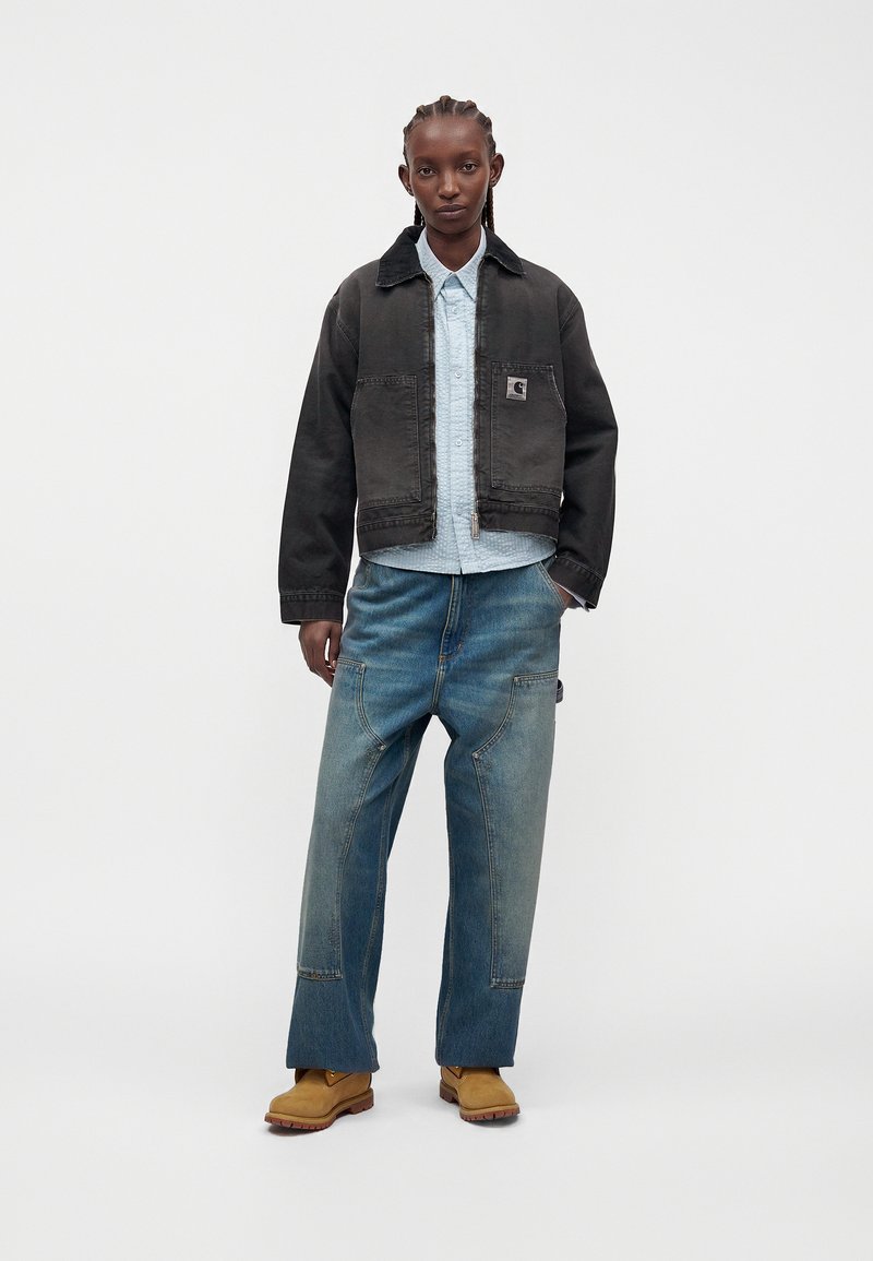 Personne aux cheveux tressés porte une veste courte foncée par-dessus une chemise bleu clair boutonnée, un jean bleu ample et des bottes de travail couleur fauve, debout sur fond blanc.