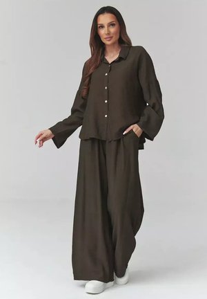 Kobieta ubrana w luźną ciemnobrązową koszulę na guziki i szerokie spodnie, stojąca z jedną ręką w kieszeni, widoczne białe trampki.