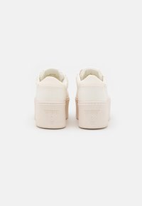 Para beżowych sneakersów na platformie z teksturowanej tkaniny i wytłoczonym logo Guess na tylnej pięcie, pokazana od tyłu na białym tle.