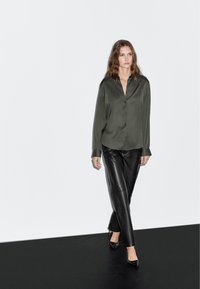 Donkergroene zijden overhemd met knopen, gecombineerd met zwarte leren broek; heeft een losse pasvorm en lange mouwen, gedragen met puntige zwarte schoenen.