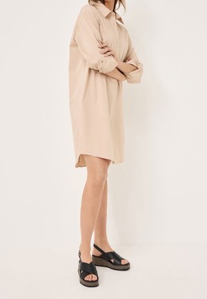 Robe chemise beige surdimensionnée avec un col pointu, des manches longues et une fente latérale. Associée à des sandales à plateforme noires à bretelles.