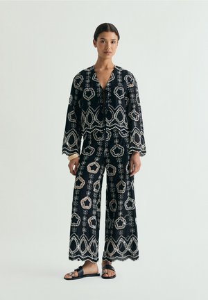 Woman wearing black wide-leg pants and matching long-sleeve top with white floral embroidery, standing on a plain light background.