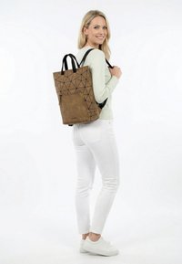 Vrouw met een lichtgroene trui, witte broek en witte sneakers, met een rugzak met een bruin geometrisch patroon op haar rug.