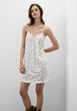 Kobieta w białej sukience bez rękawów z małymi czerwonymi kwiatowymi nadrukami i cienkimi ramiączkami, pozująca na tle jednolitej białej ściany.