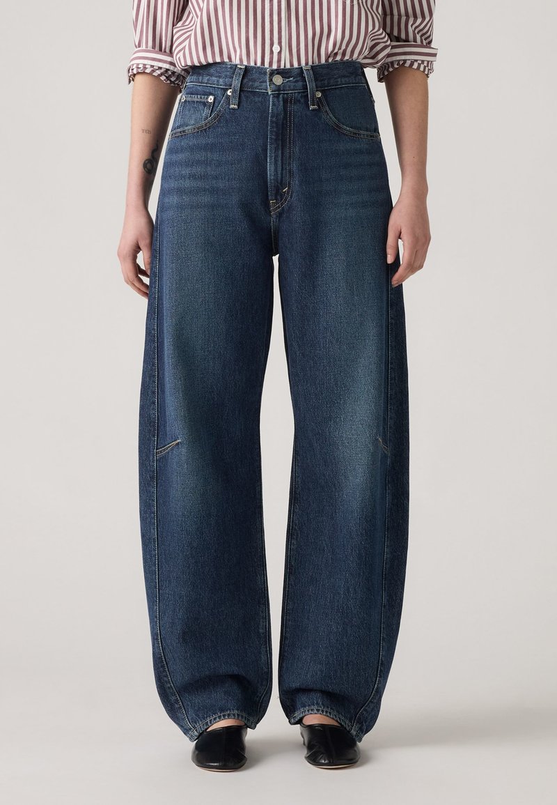Jean en denim à larges jambes de couleur bleu foncé, avec une taille haute, un léger effet délavé et des détails de couture visibles. Matériau : denim en coton robuste.