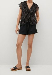 Blouse noire à volants avec imprimé floral, manches courtes, associée à un short noir et des sandales slides noires à semelles en bois.