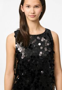 Crna majica bez rukava prekrivena okruglim šljokicama, s okruglim izrezom i glatkom teksturom, reflektira svjetlost u različitim nijansama.