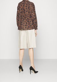 Blusa marrone con motivo floreale e colletto arricciato, abbinata a una gonna beige chiaro lungo fino al ginocchio e scarpe con tacco nere.