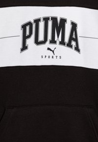 Sudadera negra con una franja horizontal blanca que presenta "PUMA SPORTS" en letras negras en negrita. Tejido suave y un detalle de bolsillo en la parte frontal.