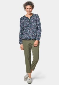 Musterbluse mit navyfarbendem Hintergrund, grünen Akzenten und langen Ärmeln, kombiniert mit olivgrünen Hosen und silbernen Schuhen.