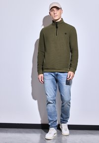 Pullover olivově zelené barvy se zipovým límcem, texturovaná látka, v kombinaci se světle modrými džíny a krémovými teniskami, na neutrálním pozadí.