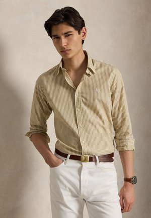 Jeune homme portant une chemise beige à rayures boutonnée, pantalon blanc, ceinture marron et montre-bracelet, debout avec une main dans la poche.