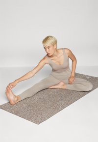 Bež yoga prostirka s teksturiranom površinom. Osoba u svijetlo bež sportskom odijelu isteže se s jednom nogom ispruženom, dosegnuvši do stopala.