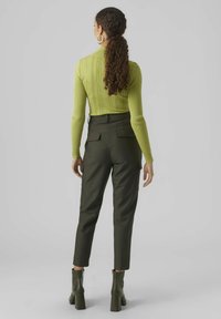 Grünes, geripptes Langarmoberteil kombiniert mit dunkelgrünen, hoch taillierten Hosen, die seitliche Taschen und einen knöchellangen Saum haben, getragen mit Stiefeln mit Absatz.