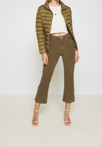 Femme portant une veste matelassée olive, un top court blanc, un pantalon en velours côtelé court olive et des talons blancs à lanières se tenant devant un fond uni.