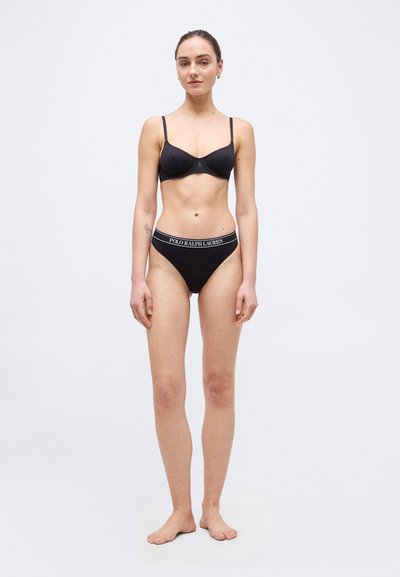 Γυναίκα, ξυπόλητη, με μαύρο σουτιέν και μαύρα, κοντά σλιπ υψηλής κοπής Polo Ralph Lauren, σε απλό ανοιχτό φόντο.