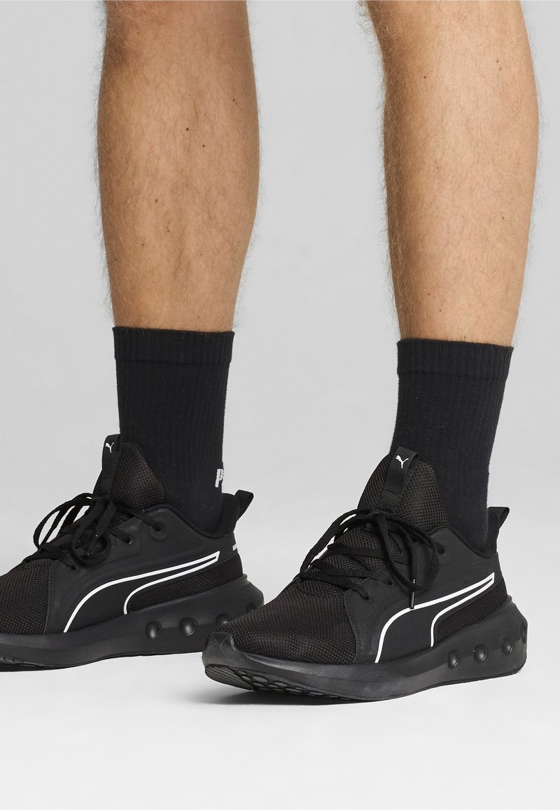 Jambes portant des baskets Puma noires avec logo blanc et chaussettes noires montantes, debout sur une surface gris clair.