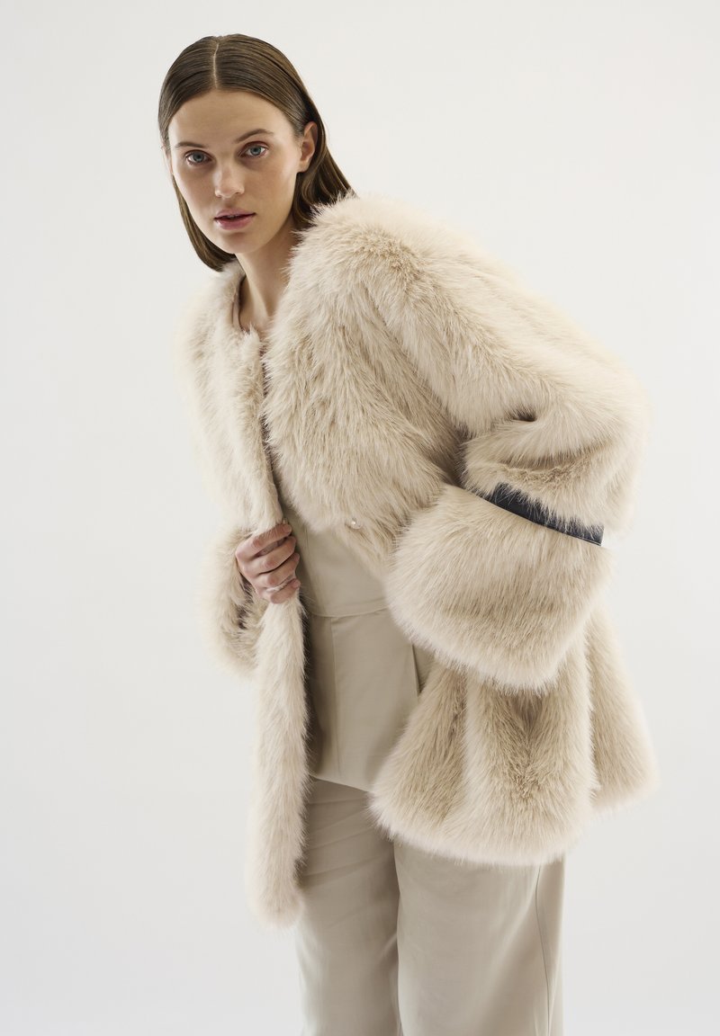 Faux-Fur-Jacke in hellem Beige mit weiten Ärmeln, ohne Kragen und einem gerippten schwarzen Akzent an den Manschetten. Glatte Textur und plüschiges Aussehen.