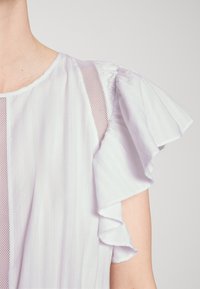 Top de tejido blanco con rayas verticales, que presenta mangas con volantes y paneles de malla. Textura ligera con un brillo sutil.