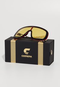 Zonnebril met een zwart frame, rode accenten en gele lenzen. Verpakt in een zwarte doos met gouden grafisch ontwerp en branding.
