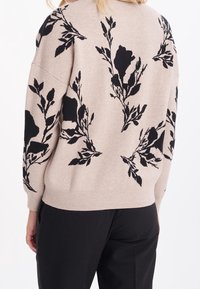 Beige tröja med svarta blommönster, med avslappnad passform, nedtagna axlar och ribbad nederkant. Bärs med svarta byxor.