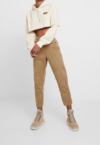 Osoba oblečená v zkráceném krémovém svetru s kapucí, béžových kalhotách s vysokým pasem a béžových vysokých šněrovacích botách, stojící na neutrálním pozadí.