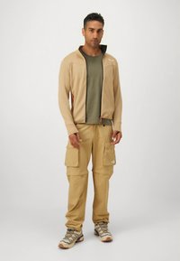 Smėlio spalvos striukė su užtrauktuku ir tekstūruota medžiaga, alyvuogių žalia marškinėliai ir chaki spalvos kelių kišenių kelnės. Šviesios spalvos žygių batai.