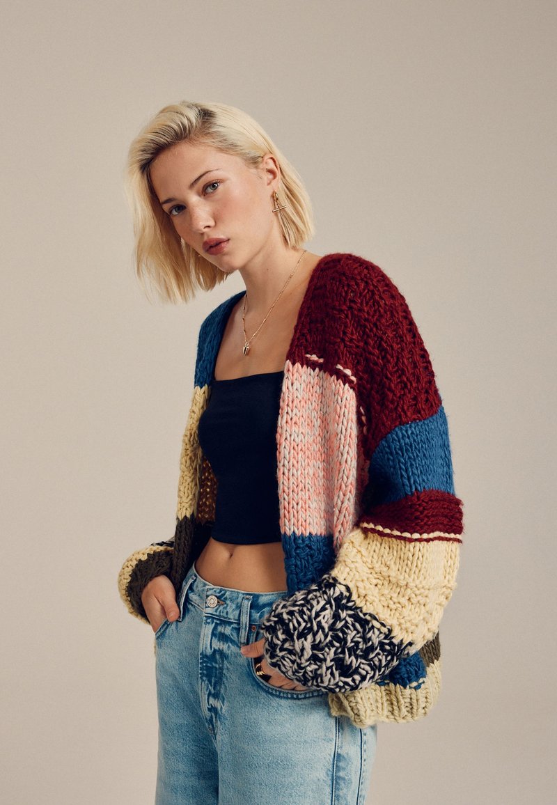 Chunky knit cardigan with multicolored patterns in red, blue, yellow, and pink. Features oversized sleeves and a relaxed fit.