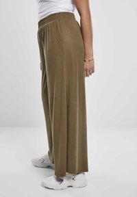 Personne portant un pantalon large olive, des baskets blanches, un haut blanc et un bracelet argenté sur un fond blanc uni.