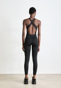 Vrouw die met haar rug naar de camera staat, gekleed in een zwarte cross-back mouwloze jumpsuit en zwarte sneakers, tegen een eenvoudige witte achtergrond.