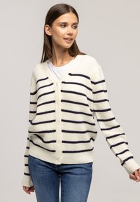 Kremowy kardigan w granatowe paski, dekolt w serek, z przodu na guziki, ściągacze przy mankietach i na dole, dzianinowa struktura. Noszony na białej koszulce, w połączeniu z niebieskimi dżinsami.