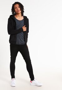 Un hoodie zippé noir sur un t-shirt gris foncé, assorti à un jean skinny noir et des baskets blanches avec des accents rouges. Texture de tissu lisse.