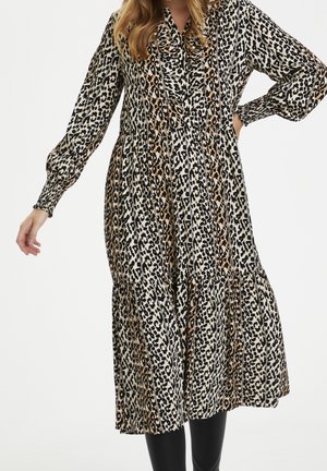 Kobieta w długiej, warstwowej sukience midi z wzorem lamparta w kolorach czarnym, białym i brązowym, w połączeniu z czarnymi legginsami.