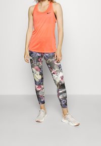 Kirjava oranssi Nike-hihaton paita yhdistettynä tiukasti istuviin kukkakuosiseksi leggingseihin, joissa on purppuran, vaaleanpunaisen ja vihreän sävyjä. Valkoiset tennarit korostuksilla.