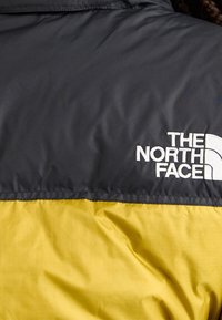 Tissu de veste noir et jaune avec le logo "The North Face" brodé en blanc sur la partie supérieure du dos.