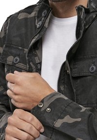 Kamouflagejacka i slitstarkt tyg, med dragkedja, knappar och flera framfickor. Grå och svart färgschema.