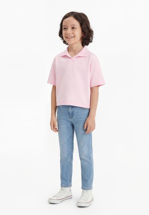 Lokkis juustega laps naeratab, kannab heleroosat polo-särki, helesiniseid teksaseid ja valgeid kõrge säärega tossusid, seisab valgel taustal.