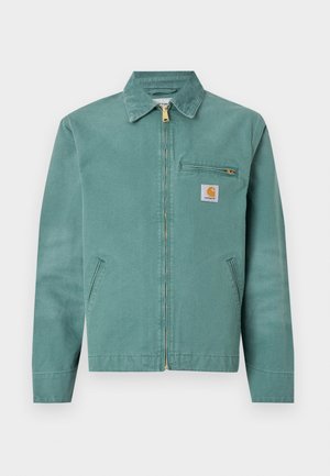 Jachetă din bumbac verde cu fermoar frontal, două buzunare laterale și un buzunar pe piept. Prezintă un patch logo pătrat cu un accent galben.