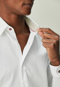 Bela srajca z teksturirano tkanino, kontrastno obrobo ovratnika z vzorcem v zeleni in rdeči barvi ter rjavimi gumbi na ovratniku in manšetah.