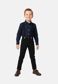 Czarne spodnie na szelkach z drewnianymi guzikami, w połączeniu z granatową koszulą zapinaną na guziki i brązowymi butami.