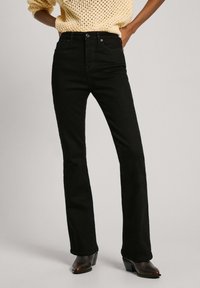 Vaqueros acampanados negros hechos de mezclilla, con cintura a media altura, bolsillos delanteros y detalles de costura sutiles. Combinados con botas marrones de tacón.