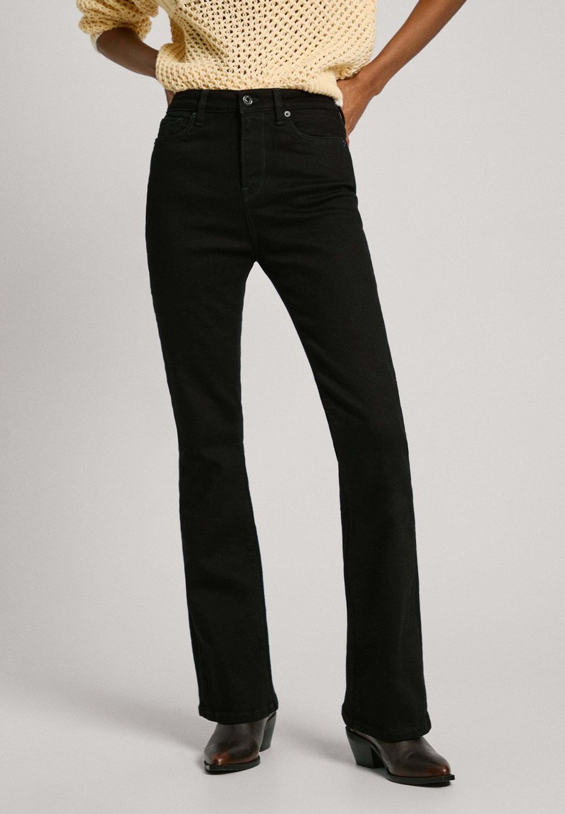 Vaqueros acampanados negros hechos de mezclilla, con cintura a media altura, bolsillos delanteros y detalles de costura sutiles. Combinados con botas marrones de tacón.